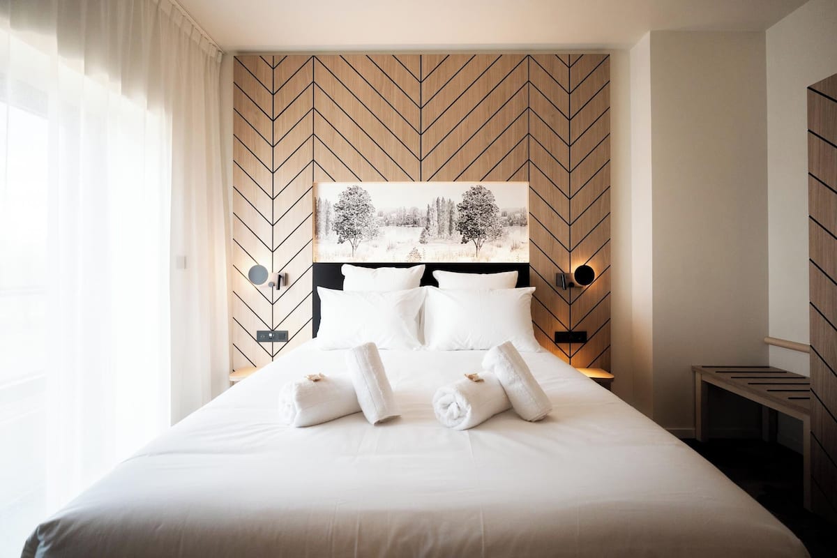 A neatly made bed with white linens and rolled towels, a large abstract painting on the wall, and a wooden headboard with a chevron pattern.