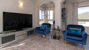 Ein Wohnzimmer mit Flachbildfernseher, zwei blauen Sesseln, einem Glastisch und einer stehenden Stehlampe.