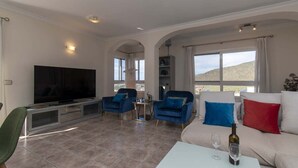 Ein Wohnzimmer mit Fernseher, zwei blaue Sessel, ein weißes Sofa mit roten und blauen Kissen und einem Tisch mit Weingläsern.