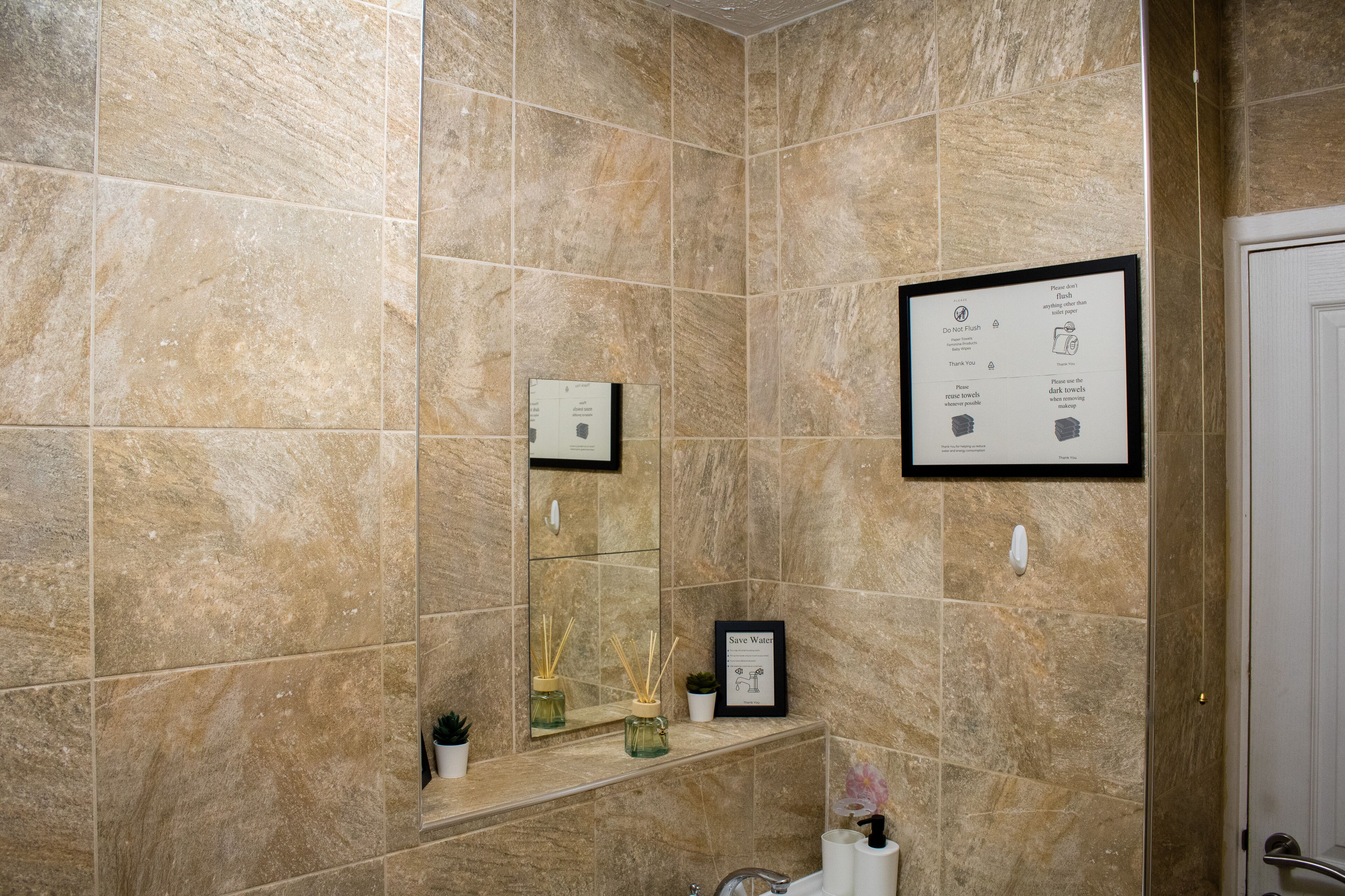 Une salle de bain avec des carreaux de marbre beige, un panneau encadré accroché au mur, un miroir, une étagère avec une plante et des toilettes.
