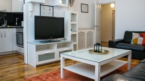 A modern living room with a flat-screen TV on a glass coffee table, a black leather sofa, and a kitchen area with white cabinetry.