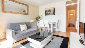 A modern living room with a grey sofa, a glass coffee table, a dining area with white chairs, and a painting on the wall. - Wandsworth Retreat: Spacious Quiet 2BR 2Bth Duplex + View + Nr Trains To Center! (London)