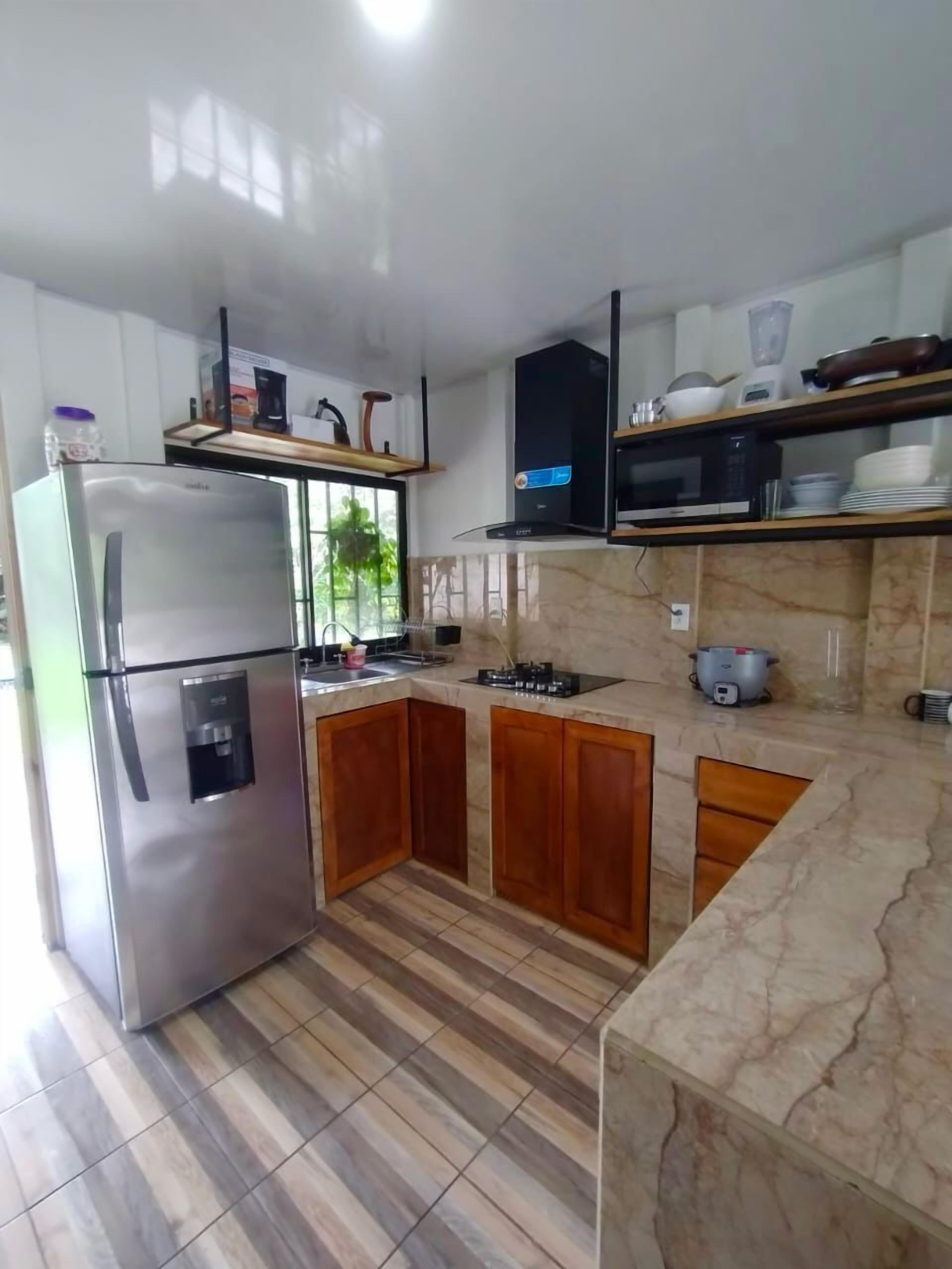 A modern kitchen with a stainless steel refrigerator, wooden cabinets, a marble countertop, and a window providing natural light.