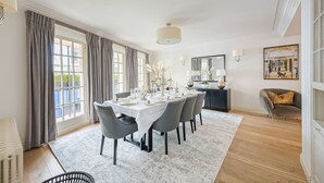 A dining room with a round table set for six, large windows with curtains, a chandelier, and a framed artwork on the wall. - Elegant 4-Bedroom London Residence in Prestigious Belgravia (London)