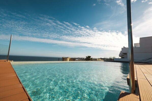 A clear pool with a wooden deck, overlooking a coastal area with buildings and the sea.