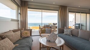 A modern living room with a sofa, a round table, and a view of the sea through large glass doors.