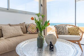 A room with a round glass coffee table, a beige sofa, and a wicker chair with a view of the sea.