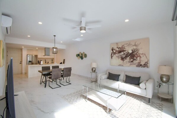 A modern living room with a white sofa, a glass coffee table, and a dining area with a kitchen in the background.