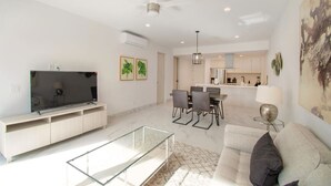 Ein modernes Wohnzimmer mit Flachbildfernseher, Glastisch, Sofa und Essbereich mit Stühlen.