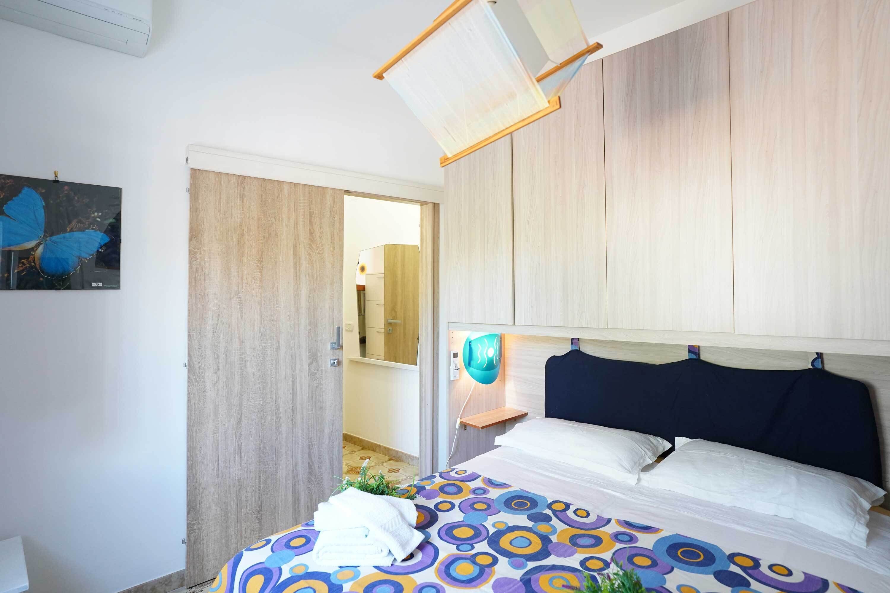 A bedroom with a bed, a nightstand, a wall-mounted lamp, and a door leading to a bathroom.