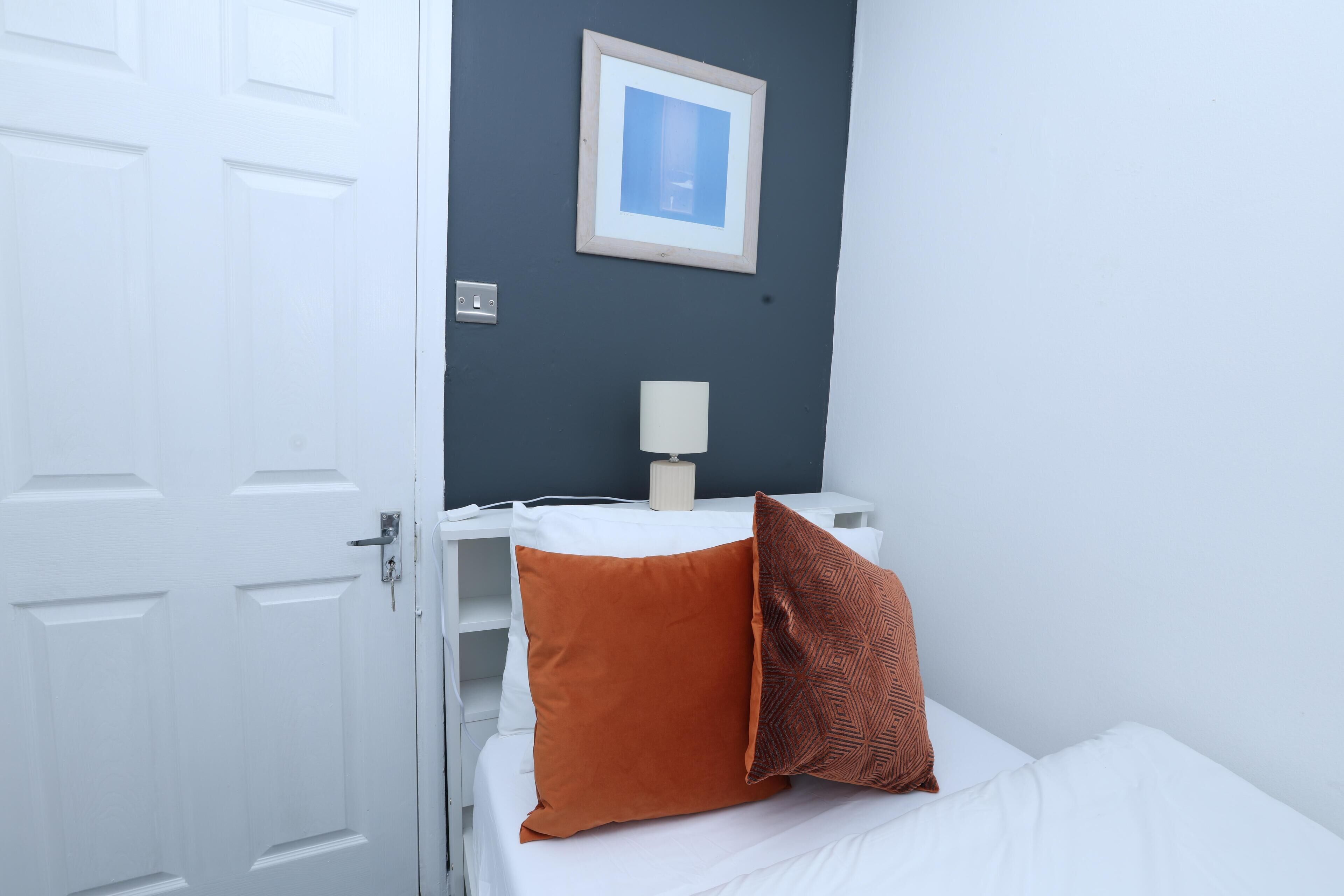 Ein Schlafzimmer mit einem weißen Bett, einem einzelnen orangefarbenen Kissen, einem gemusterten Kissen, einem weißen Nachttisch, einer Lampe und einem gerahmten Bild an der Wand.