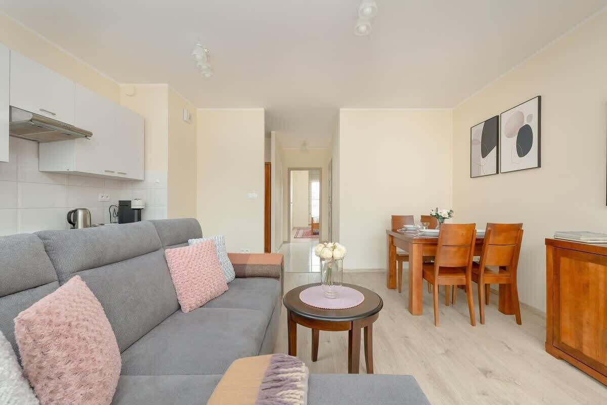 A modern living room with a grey sofa, wooden dining table, and chairs. There is a small round table with a vase of flowers. The room has a kitchenette with cabinets and a stove.