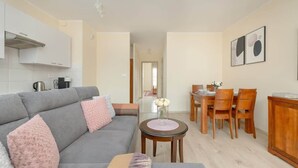 A modern living room with a grey sofa, wooden dining table, and chairs. There is a small round table with a vase of flowers. The room has a kitchenette with cabinets and a stove. - Stylish Apartment with Balcony | Guaranteed Parking (Poznan)
