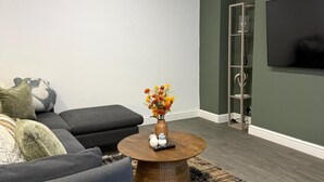 Un salon avec un canapé gris, une table basse ronde sur laquelle est posé un vase de fleurs, et un téléviseur fixé au mur.
