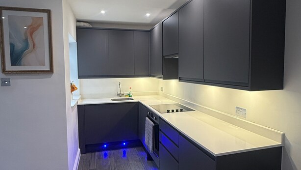 Une cuisine moderne avec des meubles de couleur sombre, un plan de travail blanc et un éclairage sous les meubles.