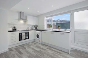 A modern kitchen with white cabinetry, a built-in oven, microwave, and washing machine, a large window with a view of the outside, and a grey wooden floor.