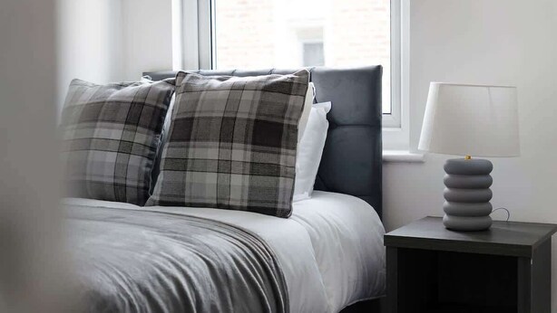 Een bed met een grijs-wit geruit kussen, een gestoffeerd grijs hoofdbord en een nachtkastje met een moderne lamp.