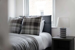 Un lit avec un oreiller à carreaux gris et blancs, une tête de lit rembourrée de couleur grise et une table de chevet avec une lampe au design moderne.