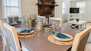 Uma mesa de jantar posta com jogos americanos, pratos e copos, com uma lareira de tijolos e uma televisão ao fundo.
