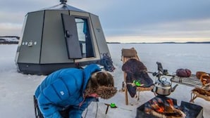 Une personne vêtue d’une veste bleue manipule un objet long sur la neige. On aperçoit également une tente et un réchaud portable avec une casserole dessus.