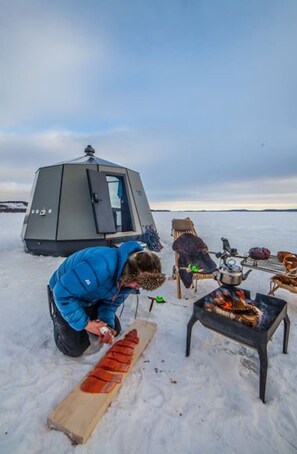 A person in a blue jacket preparing food outdoors with a portable stove and a tent in the background. - "suite With Lake View for 2 Guests Peace & Quiet Hotel in Jokkmokk Sweden" (Jokkmokk)