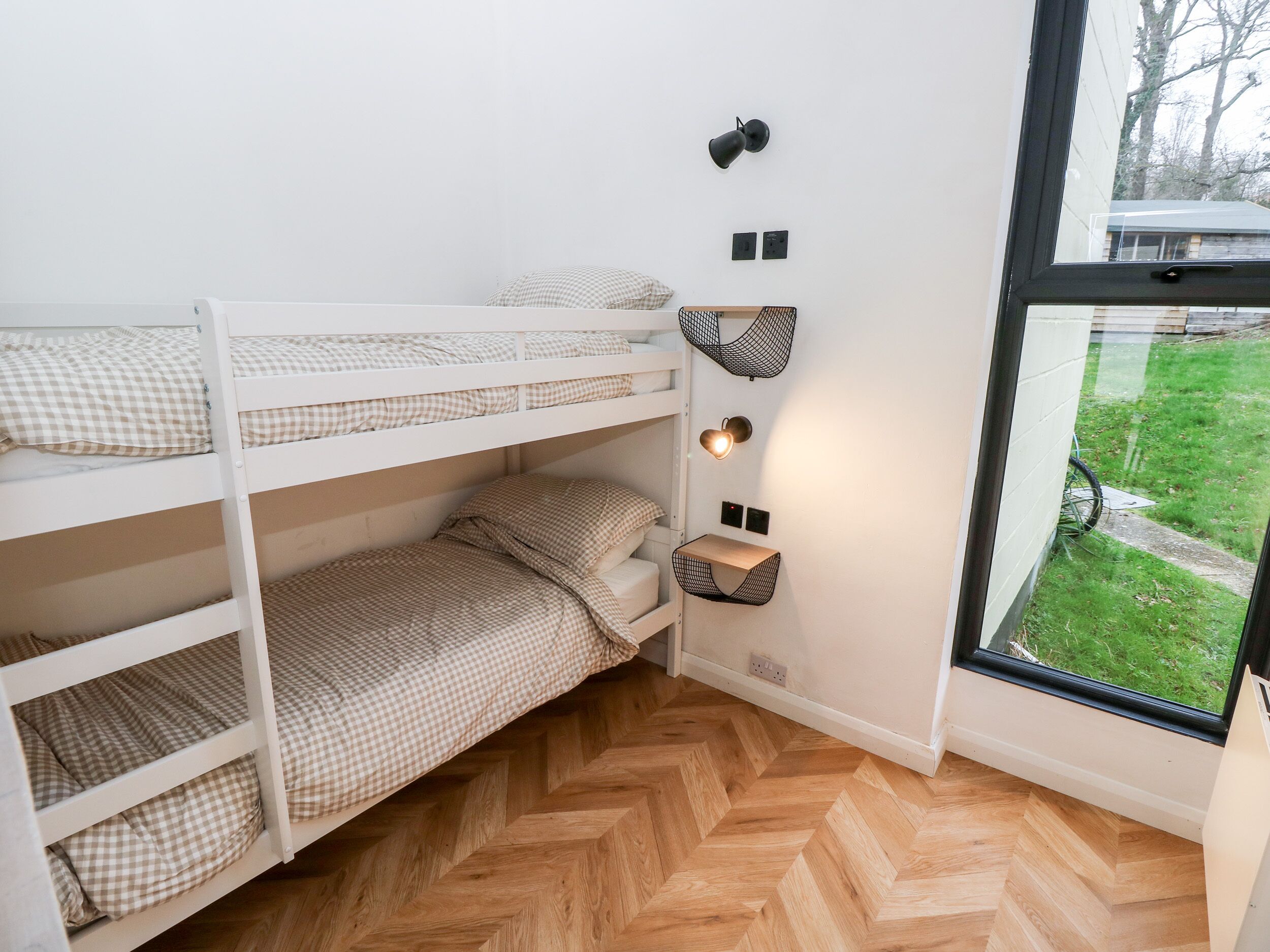A bunk bed room with a herringbone wooden floor, a window with a view of a grassy area, and wall-mounted light fixtures.