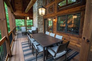 A wooden deck with a dining table and chairs, stone walls, and windows overlooking greenery.