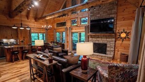 A cozy cabin interior with a fireplace, wooden walls, a dining area, and a TV mounted on the wall. - Honey Goat Hill, Sleeps 10, Centrally Located with Wooded Views (Logan)