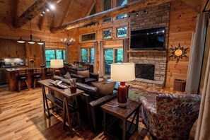 A cozy cabin interior with a fireplace, wooden walls, a dining area, and a TV mounted on the wall. - Honey Goat Hill, Sleeps 10, Centrally Located with Wooded Views (Logan)