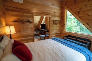 A wooden cabin bedroom with a bed, a bench, a TV, and a window with a view of trees. - Honey Goat Hill, Sleeps 10, Centrally Located with Wooded Views (Logan)