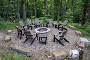Un espace de détente circulaire, avec des chaises disposées autour d’un foyer central, situé dans une zone boisée parsemée de rochers et de gravier.