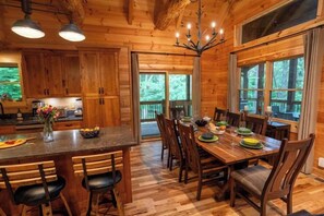 A wooden cabin interior with a dining area, kitchen, and large windows. - Honey Goat Hill, Sleeps 10, Centrally Located with Wooded Views (Logan)