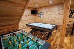 A room with a foosball table, a ping pong table, and wooden paneling.
