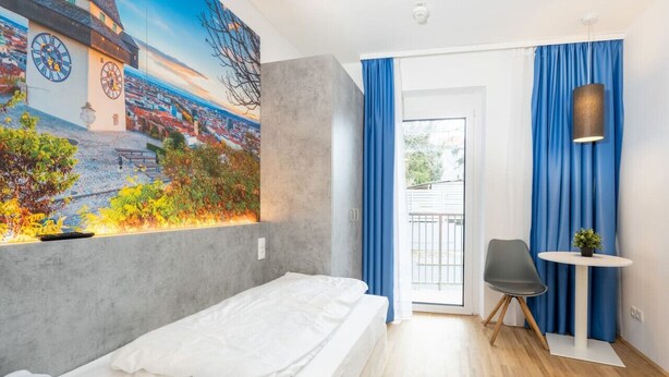 A modern hotel room with a bed, a painting of a cityscape, a balcony with blue curtains, and a small table with a plant.
