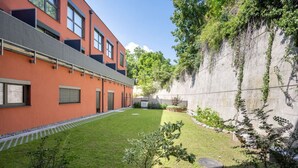 A modern building with a red facade, a small courtyard with grass, and a stone wall with greenery.