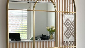 A room with a large arched mirror, a black chair, a desk with a computer, and a window with blinds.