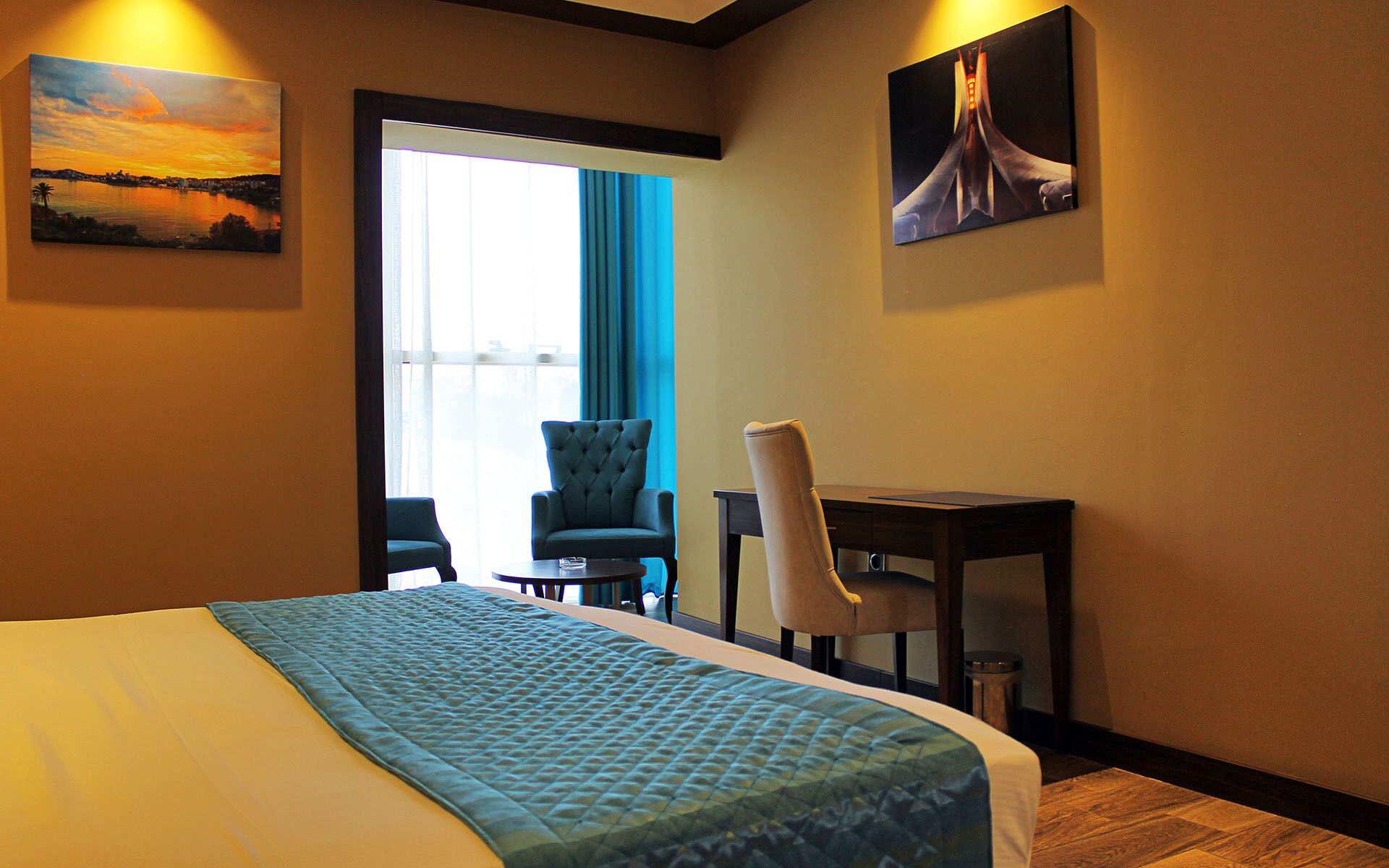 A hotel room with a bed, two chairs, a desk, and two paintings on the walls.