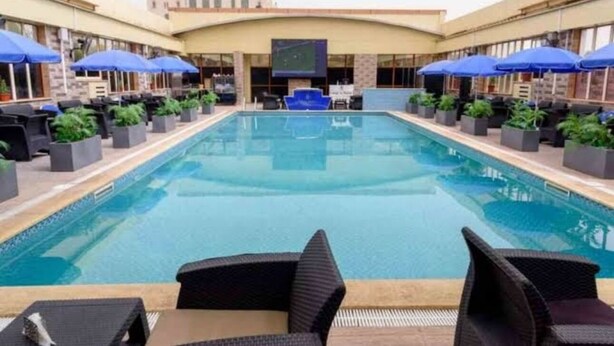 A swimming pool with lounge chairs and umbrellas, surrounded by a building with a flat roof and large windows. - LB Suites Hotel (Bir El Djir)
