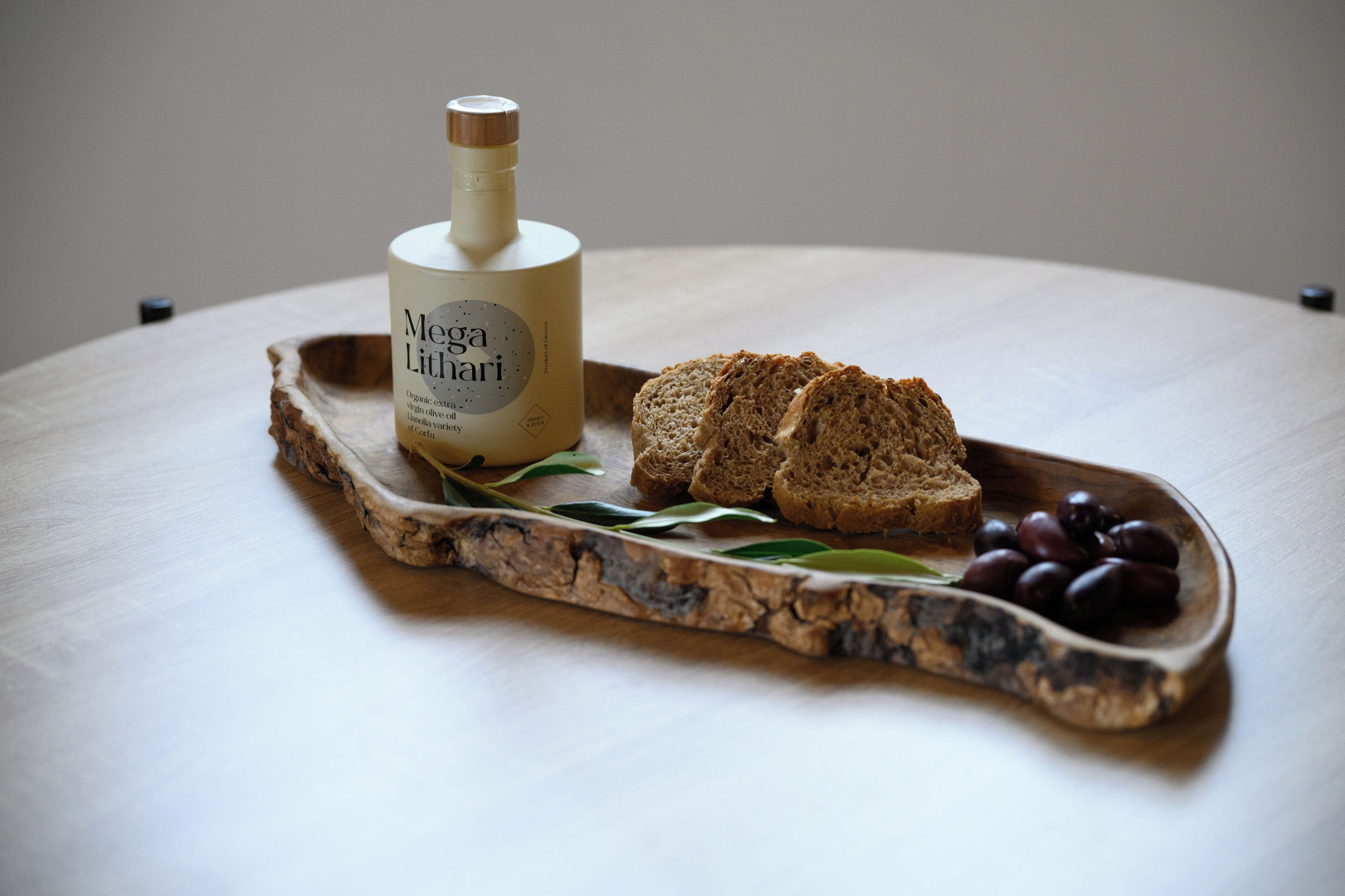 Een fles Mégá Lithárni olijfolie, een houten serveerplank met sneetjes brood en olijven.