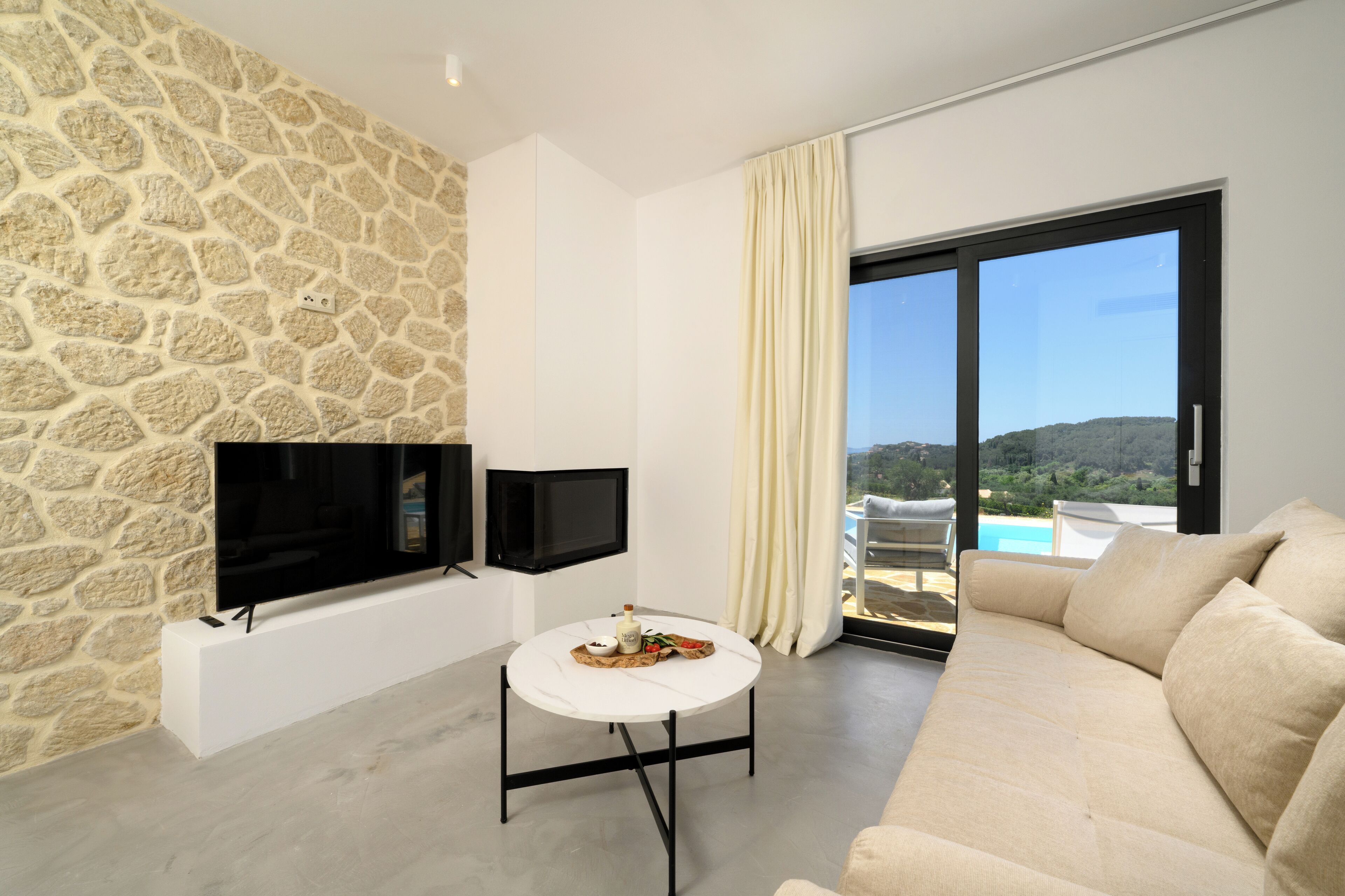 Un salon moderne avec un mur en pierre, un téléviseur à écran plat, une table ronde blanche avec une bouteille et une assiette, un canapé beige et une grande porte-fenêtre coulissante donnant sur une piscine.