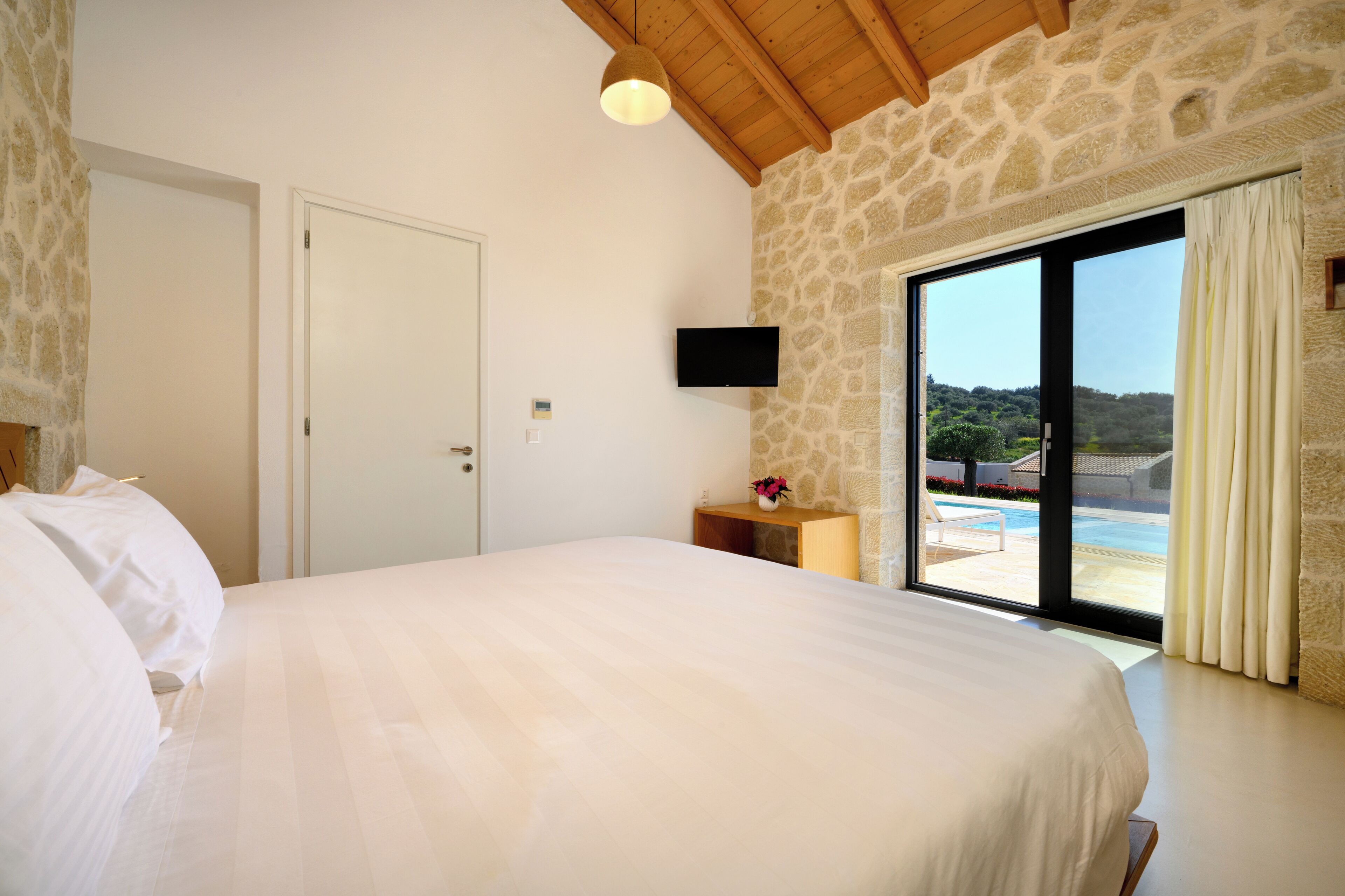 A bedroom with a large bed, a stone wall, a TV, and a view of a pool.