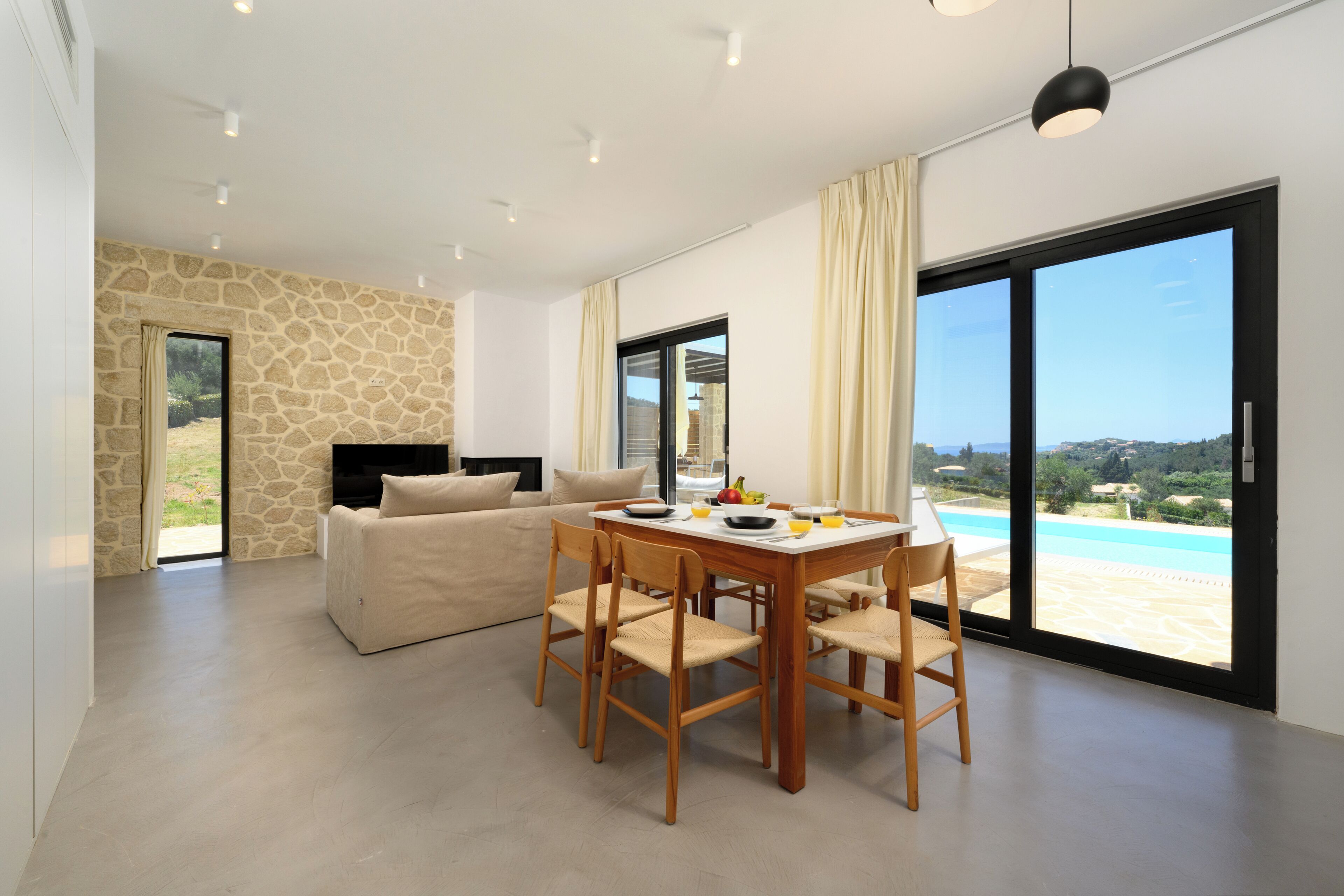A spacious living area with a stone wall, a large window with a view of a pool, a dining table with chairs, and a sofa.