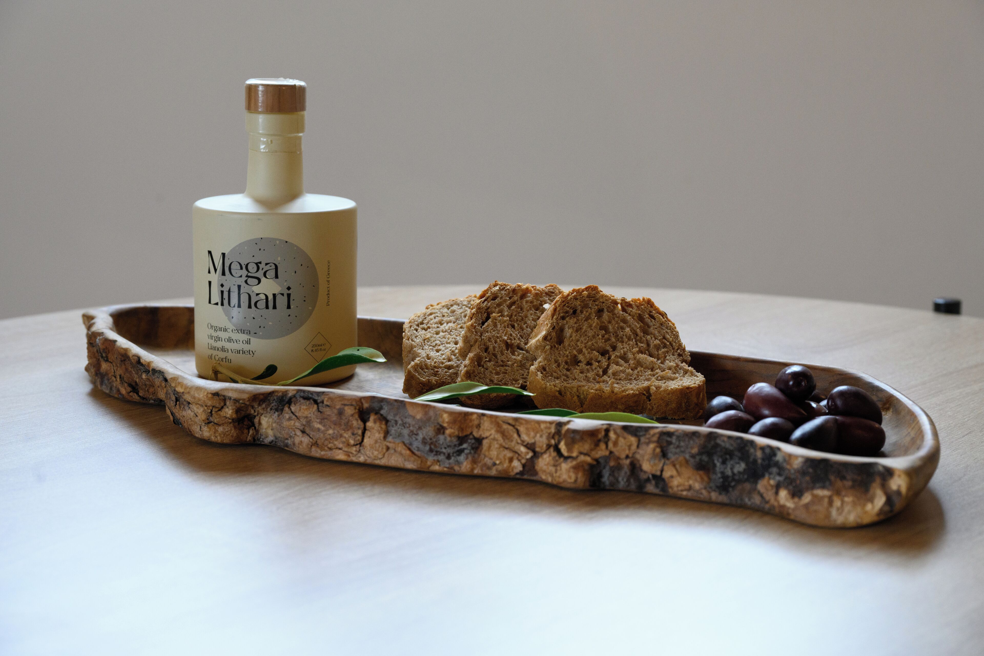Una bottiglia di olio d'oliva Mega Lithari, un vassoio di legno con fette di pane e olive.