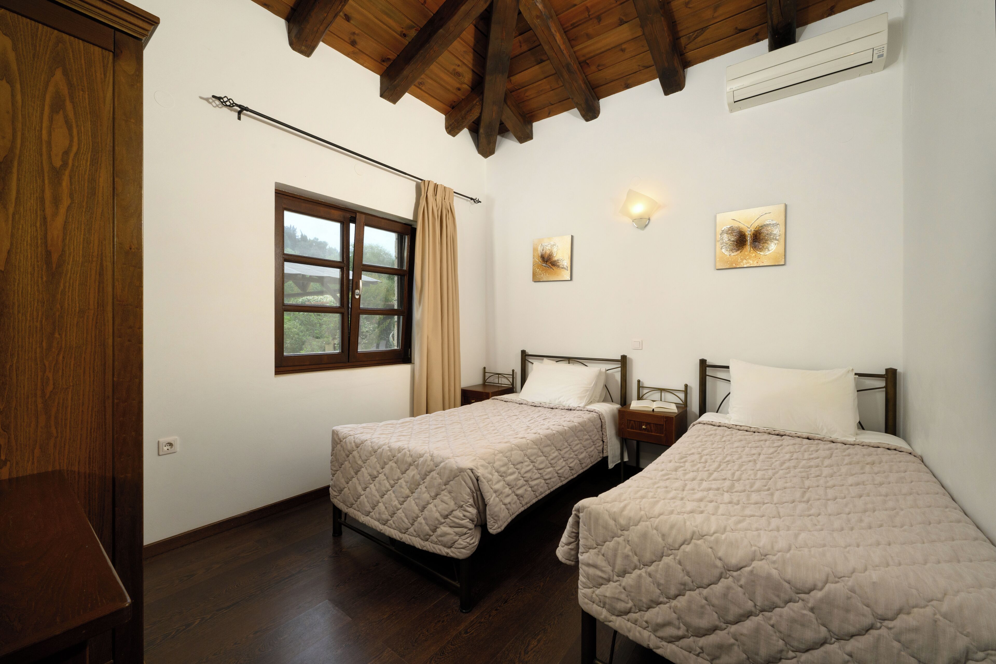 A room with two beds, wooden flooring, a window with curtains, and artwork on the walls.