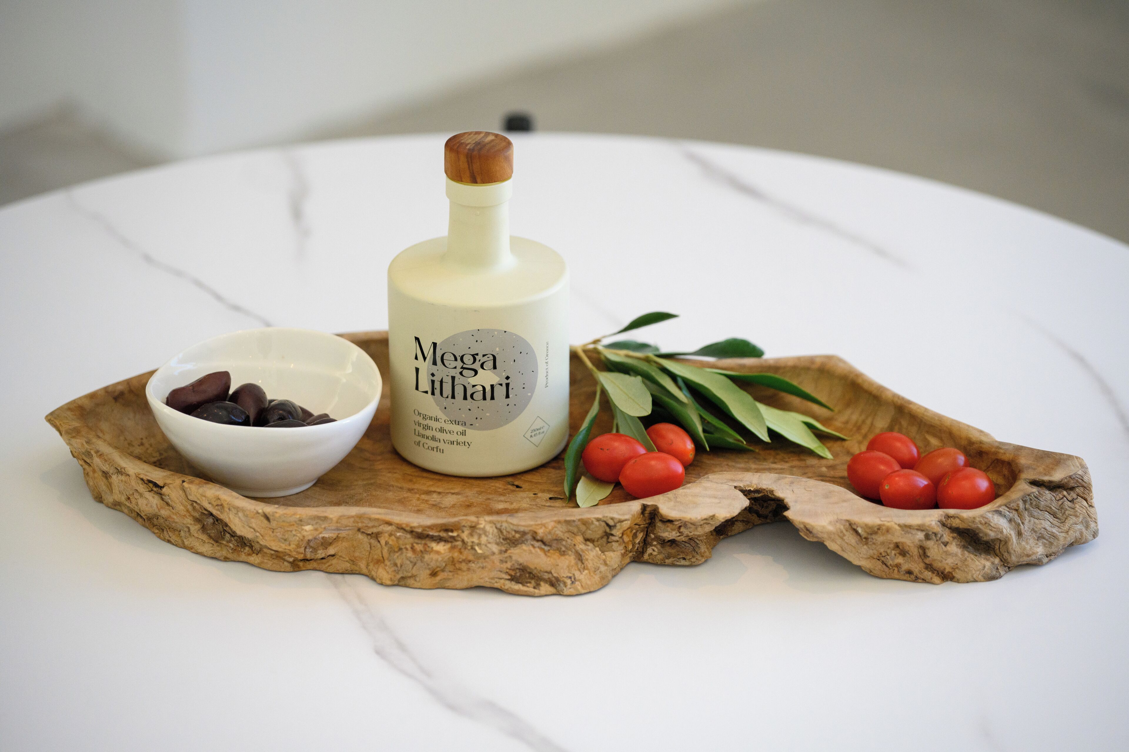 Een fles Mega Lythari biologische extra vierge olijfolie, een schaal olijven en een houten serveerplank met kerstomaatjes en groene bladeren.