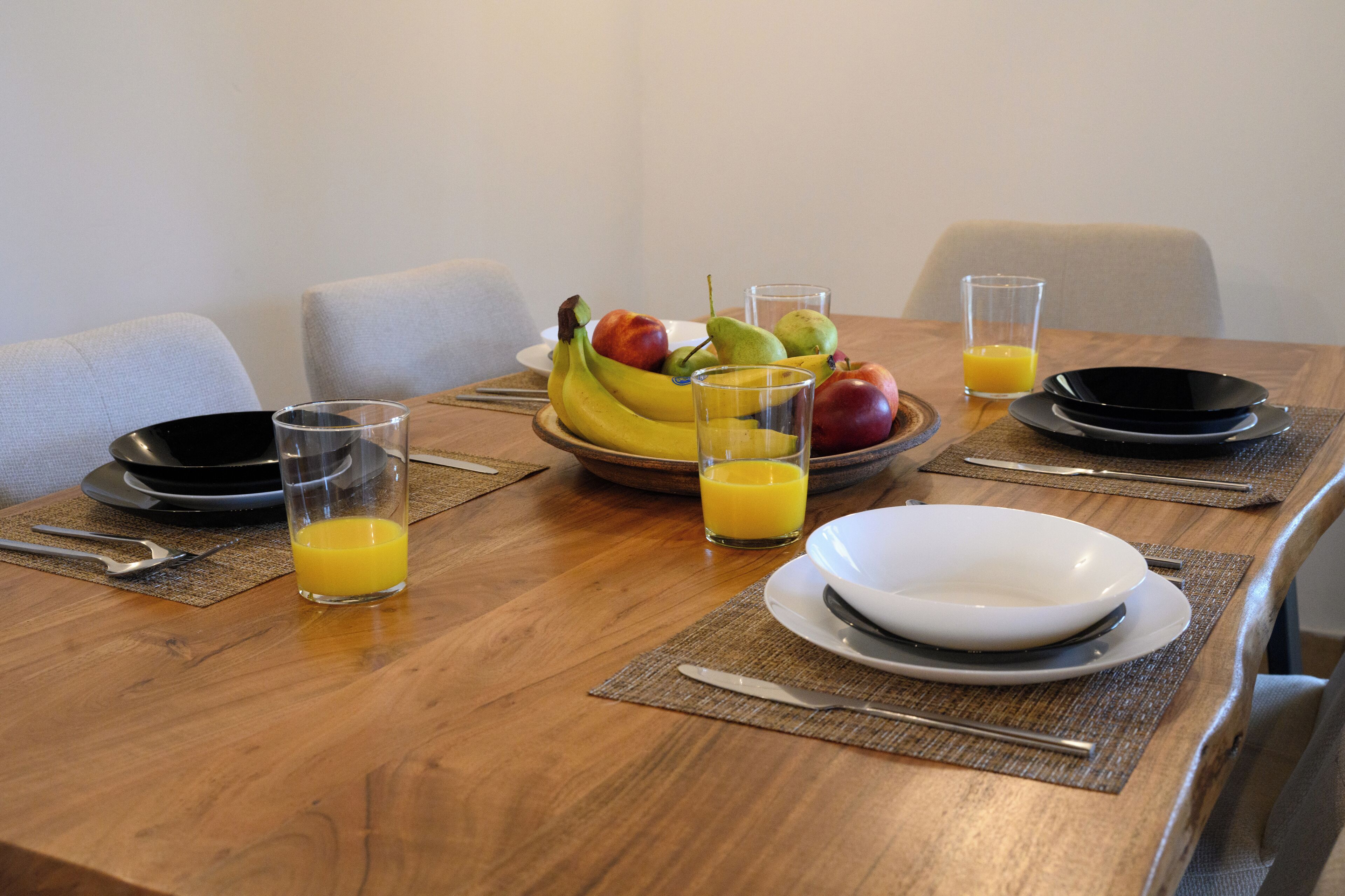 Een eettafel gedekt met borden, kommen, bestek en een fruitschaal met een glas sinaasappelsap.