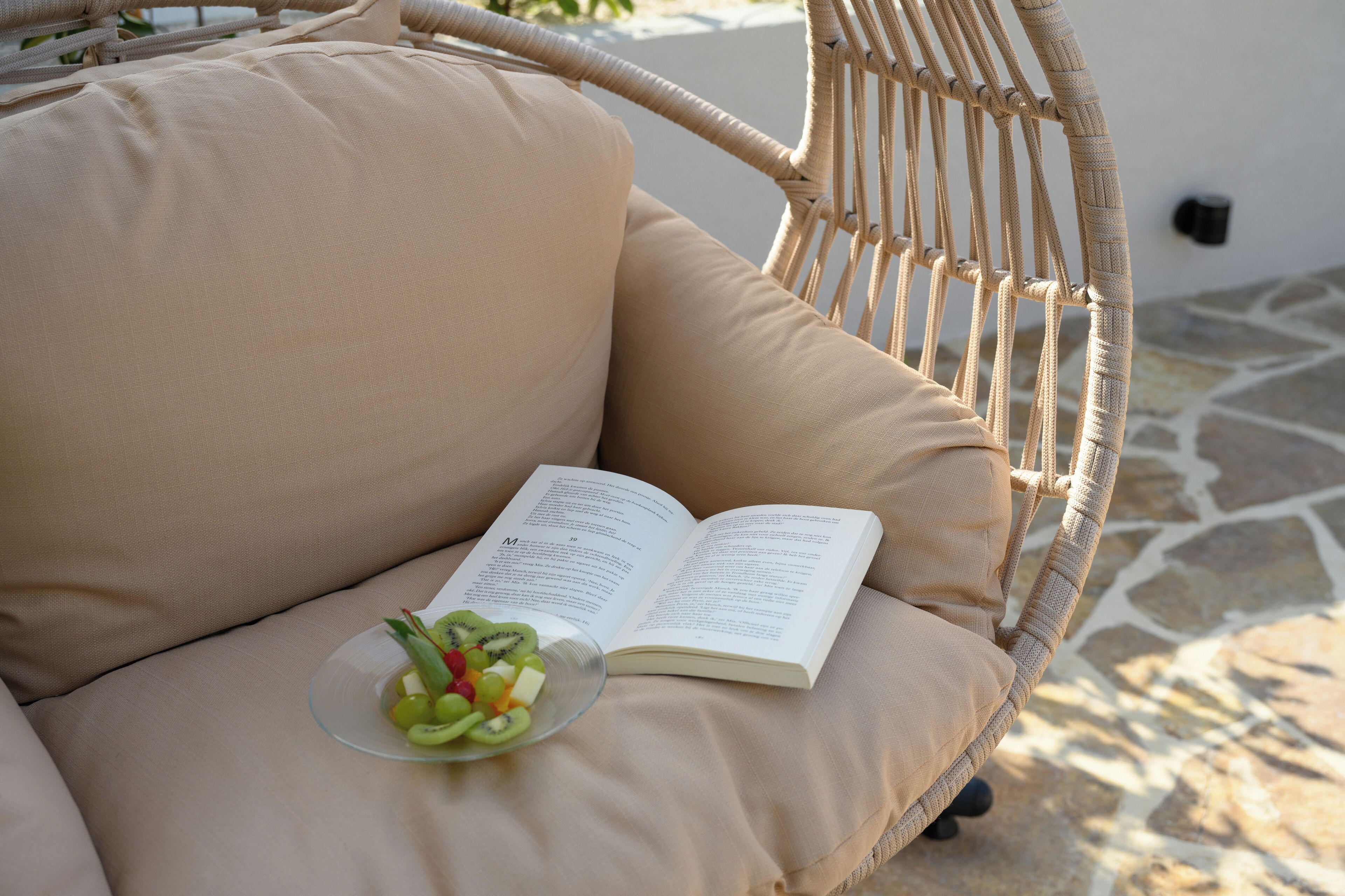 A wicker chair with beige cushions, a book, and a bowl of fruit.