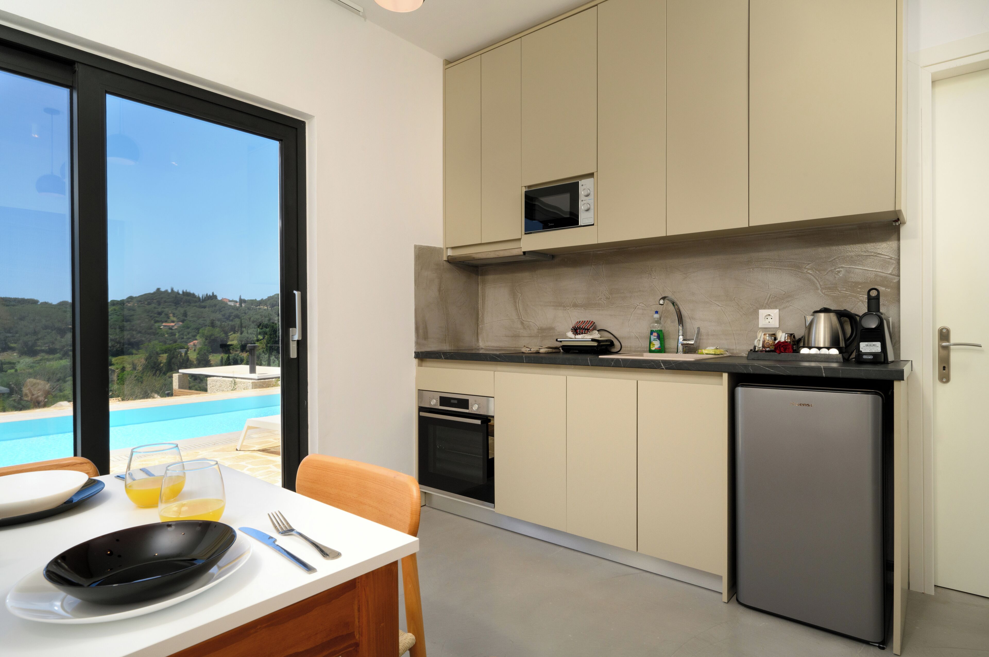 Une cuisine moderne équipée d’un four encastré, d’un four à micro-ondes et d’un réfrigérateur compact. On aperçoit une table à manger avec une assiette noire et un verre de jus d’orange.