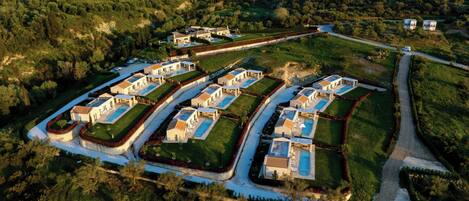 An aerial view of a residential area with multiple houses arranged in a grid pattern, surrounded by greenery and hills in the background.