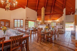 A spacious dining area with wooden tables and chairs, a stone chimney, and large windows allowing natural light. - Fazenda Realeza (Pedra Bela)